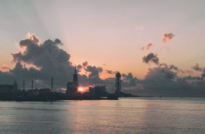 Silhouette factory by sea against sky during sunset
