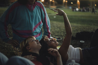 Young woman taking selfie with non-binary person while lying on friend's lap during sunset