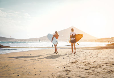 Rear view of couple holding surfboards and walking on shore of beach