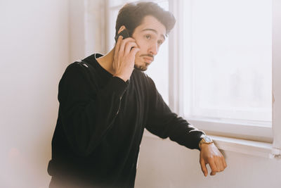 Young man talking on mobile phone by window at home