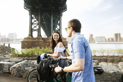 Happy couple walking with bicycle by manhattan bridge against sky