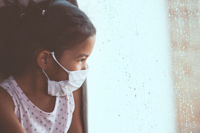 Ill girl wearing protective mask while sitting by window