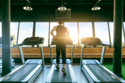 Rear view of man running on treadmill against window in gym