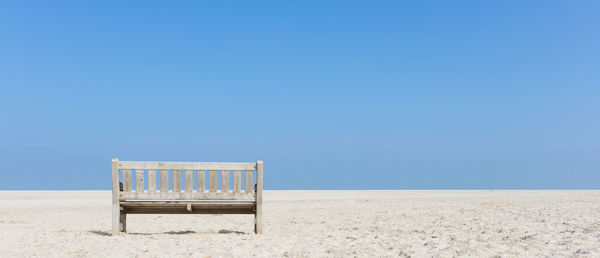 Empty bench on sand against clear blue sky