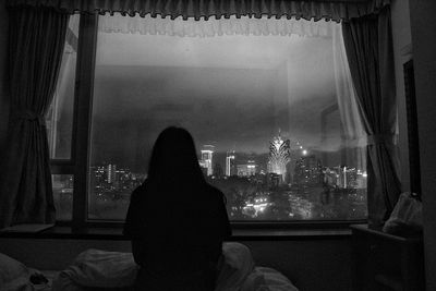 Rear view of silhouette woman looking at city through window at home