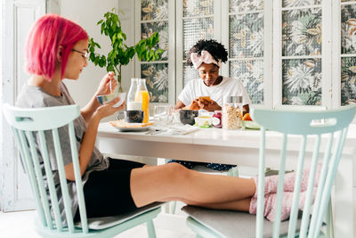 Relaxed young woman with dyed hair sitting near table and eating yummy dish near african american friend spilling cereals during breakfast in cozy apartment