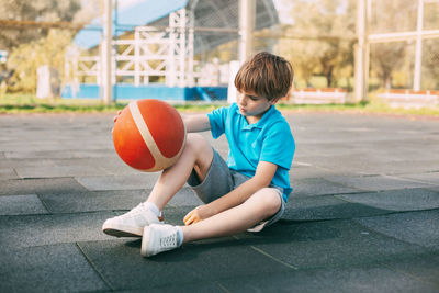 A boy basketball player in a blue t-shirt is sitting on the basketball court with a ball in his hand