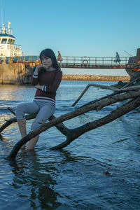 Portrait of teenage girl sitting on branch in river against clear sky