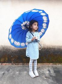 Thoughtful girl holding blue umbrella while standing against wall