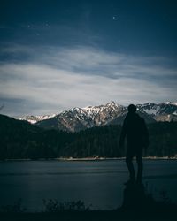 Rear view of man standing by lake against mountains and sky