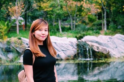 Portrait of young woman smiling while standing by pond at park