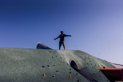 Low angle view of man standing on outdoor play equipment against clear blue sky