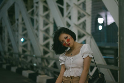 Portrait of young woman with head cocked wearing sunglasses standing on bridge in city at night