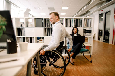 Disabled male professional sitting on wheelchair while smiling businesswoman using laptop at work place