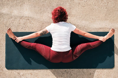 From above top view back view of anonymous barefooted woman in sportswear doing yoga in wide angle seated forward bend pose on mat training alone on street