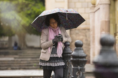 Woman holding umbrella while using phone by railing in city