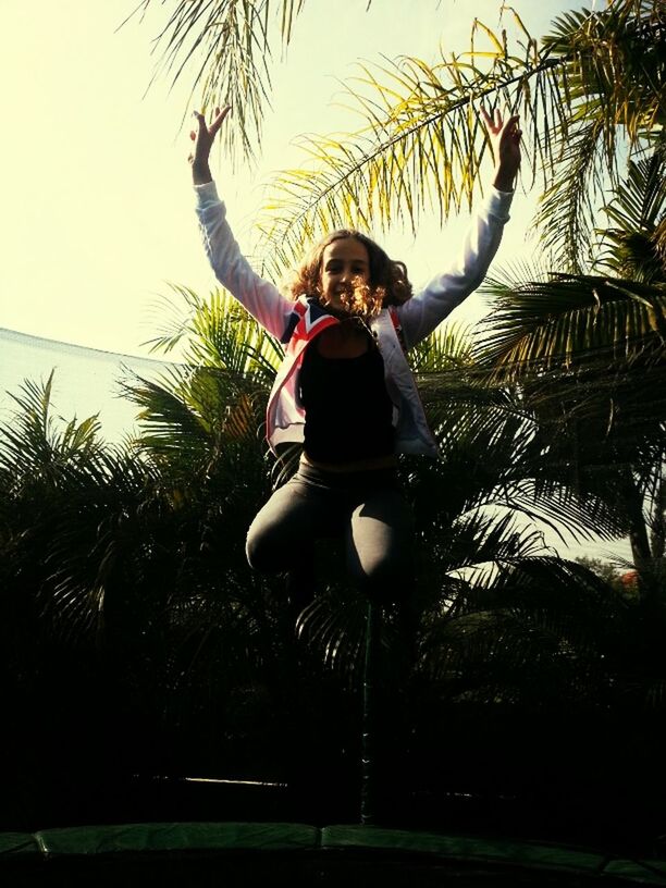 young adult, lifestyles, leisure activity, tree, full length, low angle view, looking at camera, person, young women, front view, portrait, mid-air, casual clothing, palm tree, fun, standing, sky