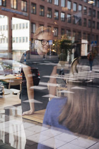 Smiling young transgender barista talking with female customer seen through cafe window