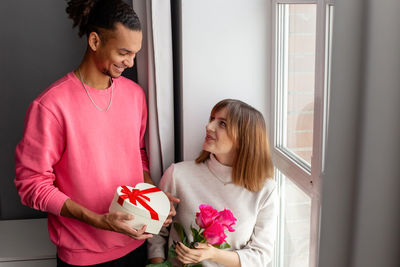 A guy in a pink jumper gives his girlfriend, a white heart-shaped box and a bouquet of roses. 