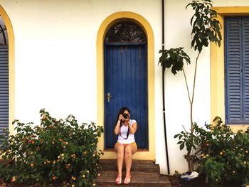Full length of young woman photographing while sitting against closed blue door