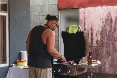 Back view of ethnic male in apron placing raw marinated meat on grill of barbecue cooker while cooking steaks