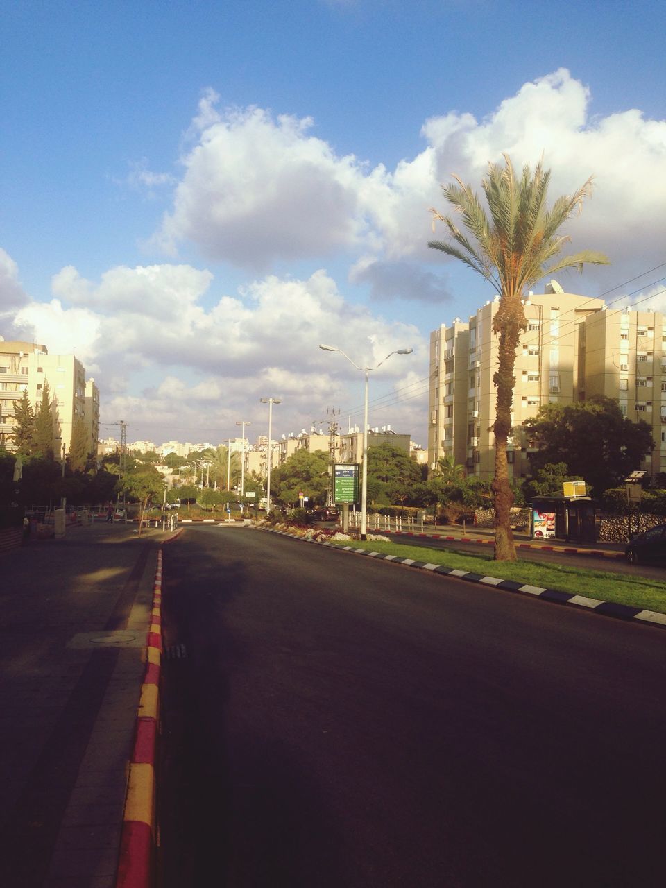 sky, building exterior, the way forward, tree, architecture, built structure, cloud - sky, road, transportation, city, road marking, diminishing perspective, street, cloud, vanishing point, palm tree, cloudy, outdoors, growth, day