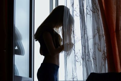 Side view of woman standing by curtains at window