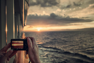 Cropped hand photographing sea from boat during sunset