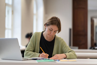 Middle-aged female student preparing for exam in public library while sitting at table with laptop