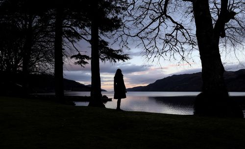 Silhouette woman standing on lakeshore against sky during sunset