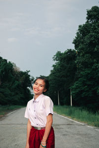 Portrait of smiling young woman standing on road against trees
