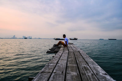 Side view of man sitting on jetty in sea against sky