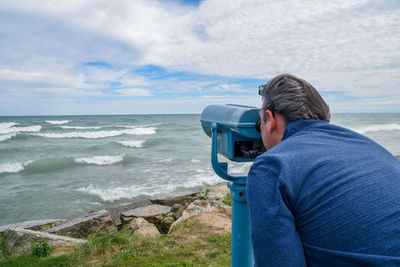 Rear view of man looking through coin-operated binoculars at lakeshore against cloudy sky