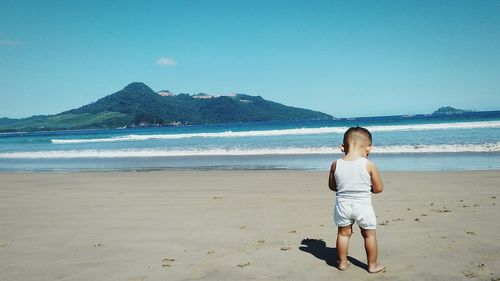 Rear view of baby boy standing on shore at beach against sky