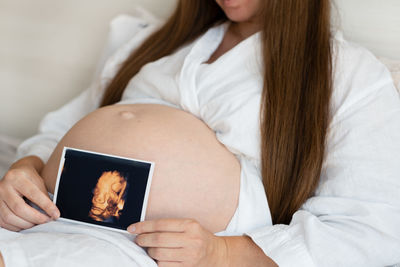 Midsection of pregnant woman lying on bed holding ultrasound photo 