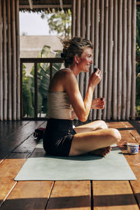 Side view of smiling woman smelling oil while sitting on yoga mat at wellness resort