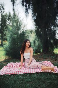 Portrait of beautiful young woman sitting on picnic blanket against trees in forest