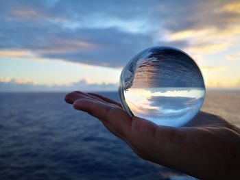 Cropped image of hand holding crystal ball against sea during sunset