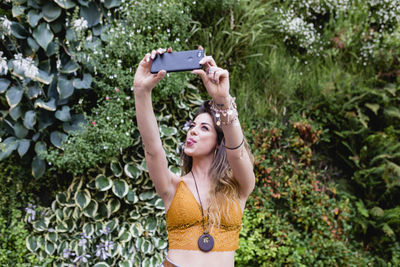 Beautiful woman taking selfie from mobile phone while standing against plants at park