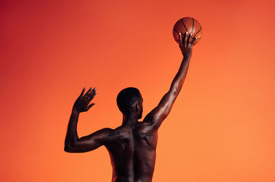 Rear view of shirtless man with arms raised against orange background