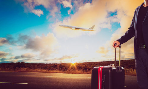 Midsection of man with luggage standing on road against cloudy sky during sunset