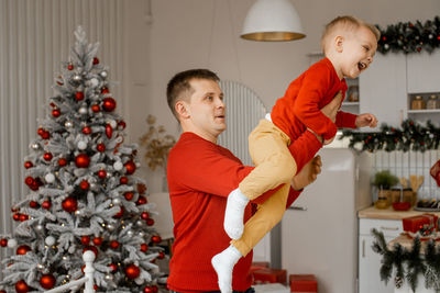 Playful father in red sweater lifting his laughing happy son in his arms by the christmas tree