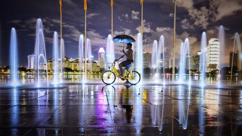 Man cycling by illuminated fountain in city at night