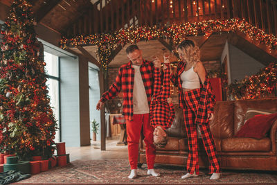 Smile active parents with small son in red checkered sleepwears waiting santa indoor.