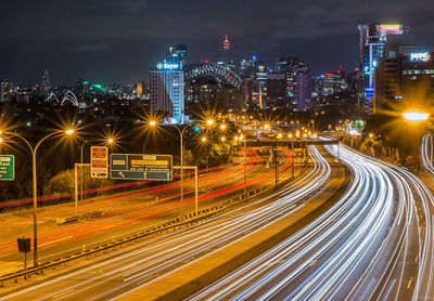 Light trails on road by illuminated buildings against sky at night