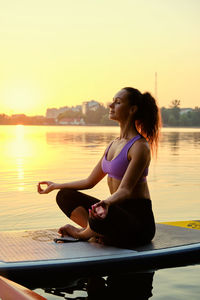 Woman sitting in sea against sky during sunset