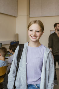 Portrait of smiling female student carrying backpack while standing in classroom at school