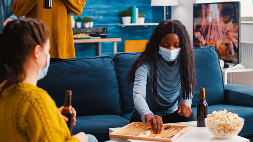 Women wearing mask playing game while sitting on sofa at home