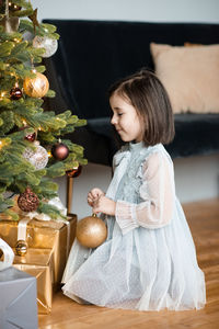 Happy little girl 5-6 year old holding christmas balls decorating tree in room. 