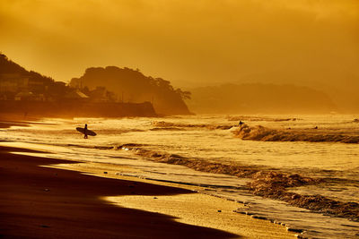 Scenic view of sea with silhouette surfer standing against orange sky during sunrise